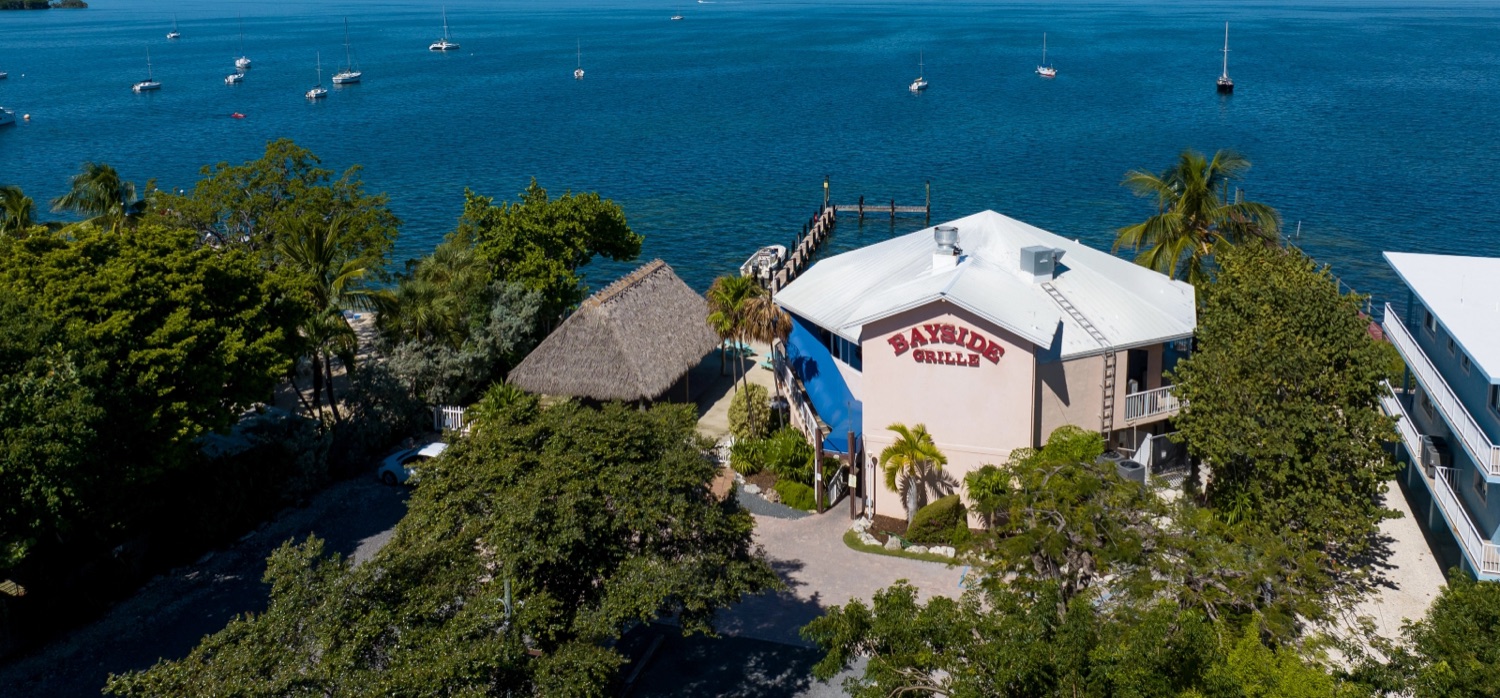 Aerial view of Bayside Grille on the shore of Florida Bay in Key Largo, with sailboats moored offshore and a beachfront tiki hut