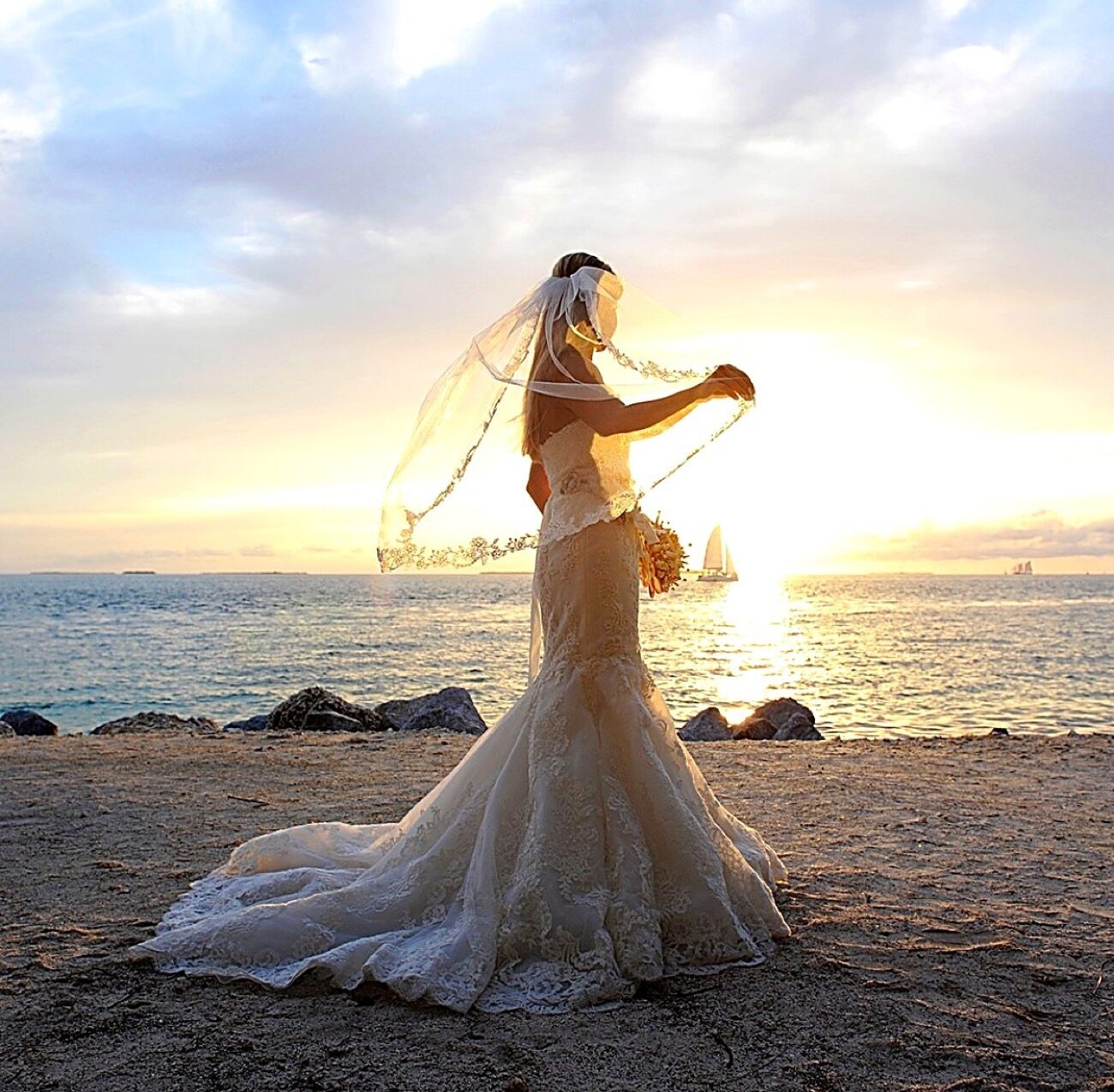 A bride in a lace gown and veil on a Key Largo beach at sunset with a sailboat on the horizon