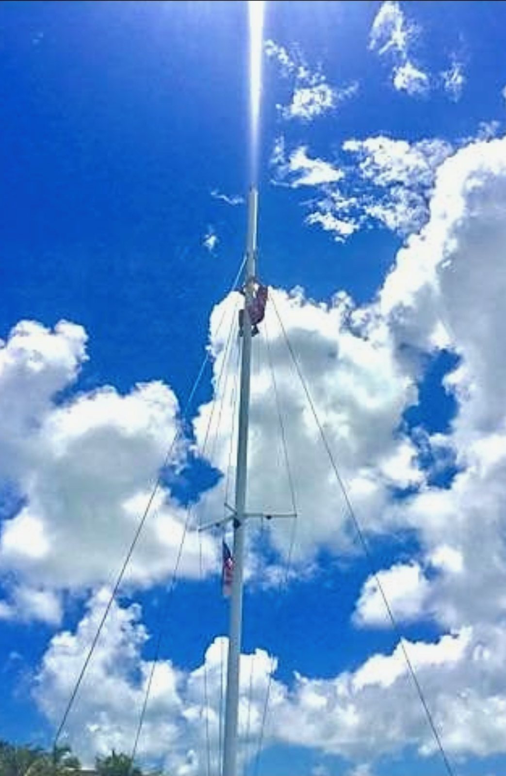 A crew member climbing the mast of the Carolina Moon catamaran against a bright blue sky with clouds