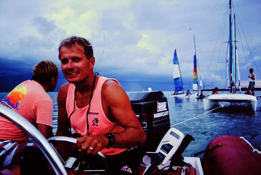 A young man in a pink shirt at the helm of a boat with Hobie catamarans sailing behind him