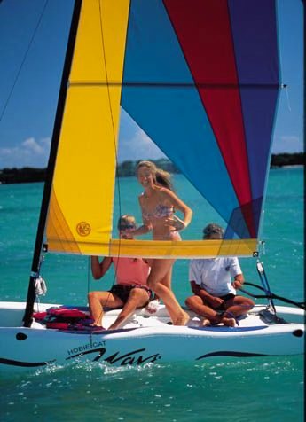 Guests laughing aboard a Hobie Wave catamaran with yellow and red sails on turquoise water