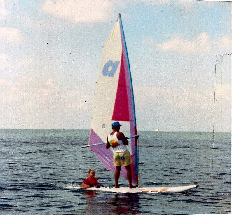 A parent windsurfing with a small child sitting on the front of the board