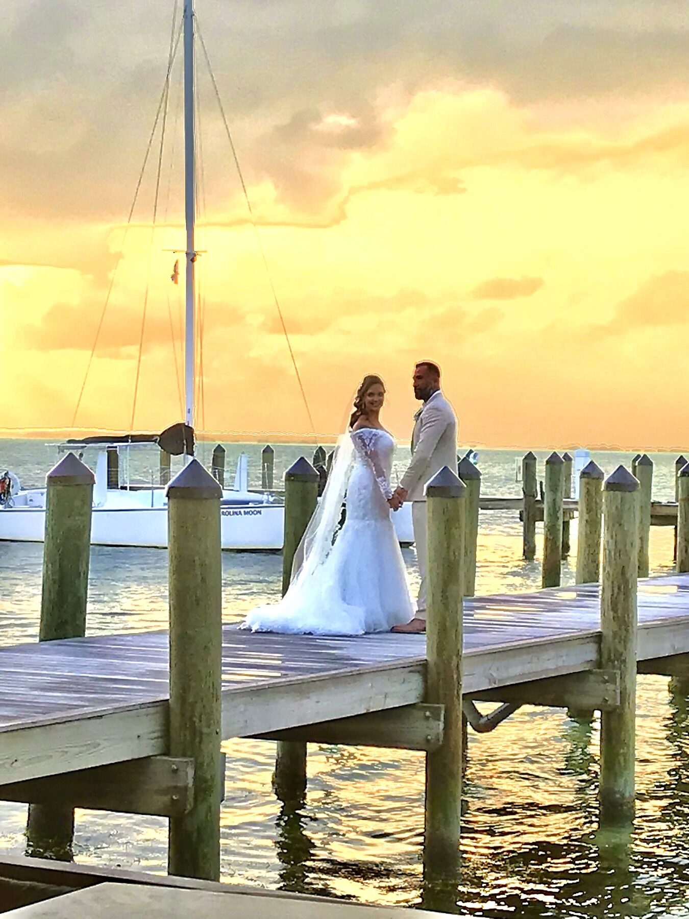 A bride and groom holding hands on a Key Largo dock at golden hour with the Carolina Moon catamaran behind them